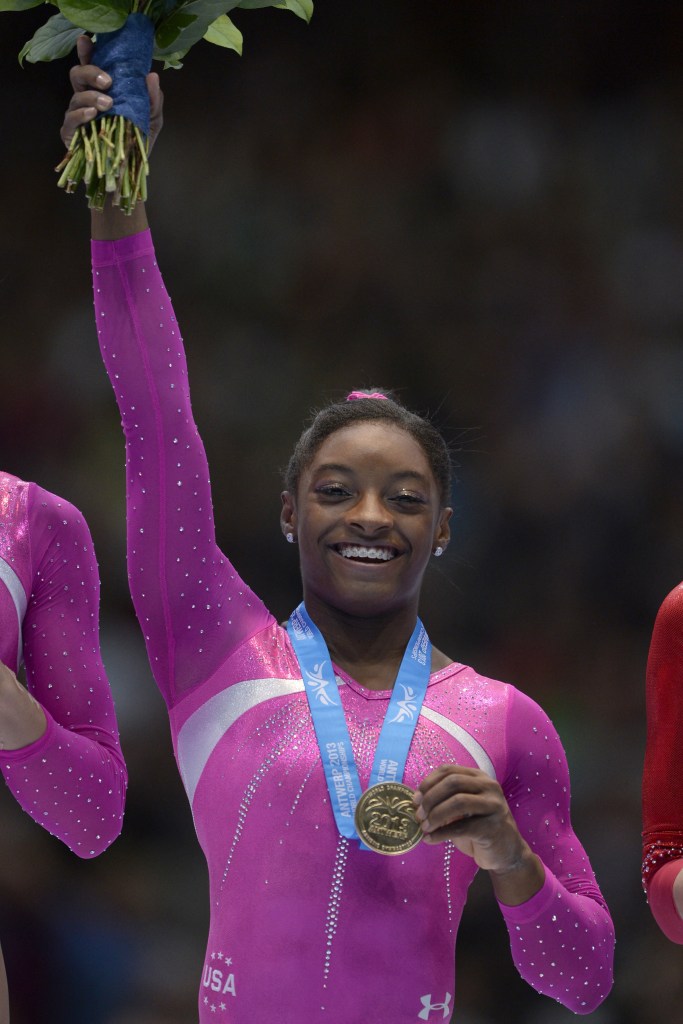 Simone Biles celebrates on the podium by holding up her flowers and grabbing the gold medal around her neck.