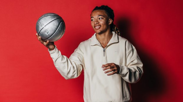 Kingston Flemings looks at a silver basketball in his hand while smiling in front of a red background.