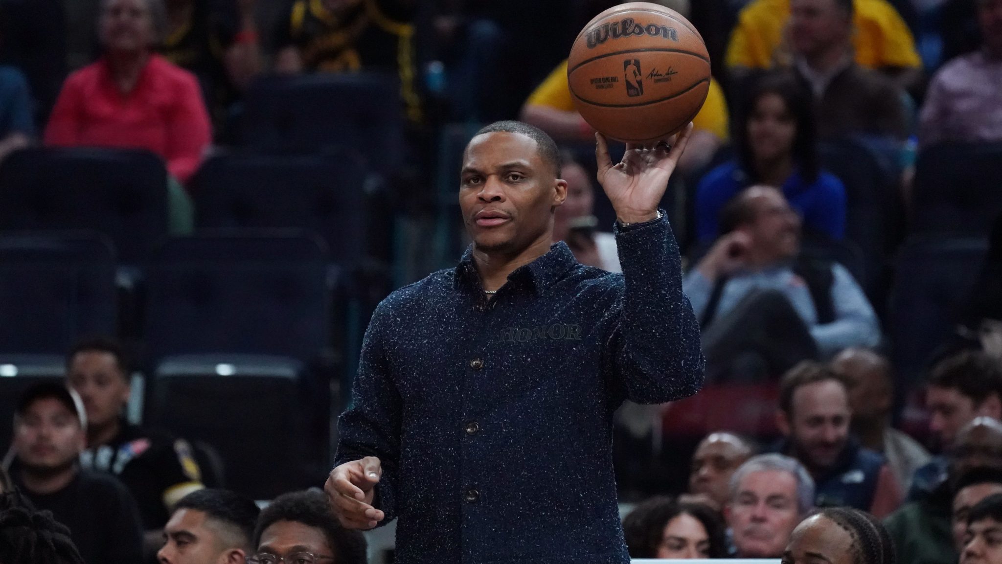 Russell Westbrook holds a basketball up with one arm on the sidelines.